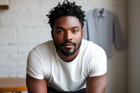 Young African American man with curly hair and beard wearing white t-shirt smiling confidently at camera against light brick wall background. Professional headshot style portrait.