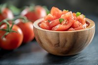 Fresh chopped tomatoes in wooden bowl with whole tomatoes on vine against dark background, close up view for cooking and food photography.