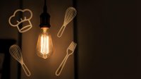 Vintage Edison bulb illuminating wooden kitchen utensils and chef hat silhouette on dark background for culinary creativity and cooking inspiration.