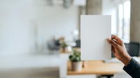 Person of color holding blank white paper sheet in modern office workspace with natural light and blurred background for mockup and branding purposes.