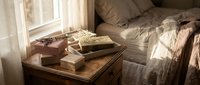Cozy bedroom corner with wooden nightstand, decorative boxes, dried lavender, and soft linen bedding in warm sunlight by a sheer curtain.
