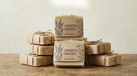 Handmade soap bars with lavender and chamomile labels stacked in pyramid arrangement on wooden surface against neutral background. Suitable for wellness, beauty, spa product marketing.