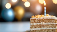 Slice of layered cake with cream frosting and single lit candle on top, celebrating special occasion against blurred festive bokeh background.