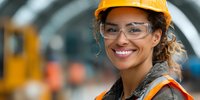 Happy female construction worker wearing safety helmet and glasses smiling at camera in industrial warehouse setting with machinery background.