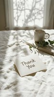 Thank you card on white linen bed with ceramic mug, books, and eucalyptus branch in soft morning sunlight near window.