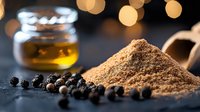 Ground black pepper powder and whole peppercorns on dark stone surface with glass jar of olive oil and warm bokeh lights in background.