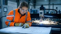Female engineer in orange safety gear welding metal in industrial factory workshop, reviewing technical blueprints and plans for manufacturing production work.