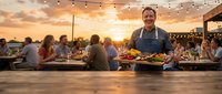 Smiling chef presenting grilled dishes at outdoor restaurant during golden hour sunset with string lights and dining guests in background.
