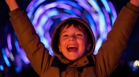 Young Caucasian boy with arms raised celebrating joyfully under colorful neon light tunnel creating vibrant purple and blue illuminated background for entertainment concepts.