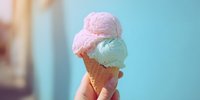 Hand holding waffle cone with two scoops of pastel ice cream, pink and mint green, against soft turquoise background, perfect for summer dessert promotions.