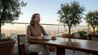 Young professional woman enjoying morning coffee on rooftop terrace with city skyline view and olive trees creating peaceful urban lifestyle moment.