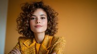 Young woman with curly brown hair and fair complexion wearing yellow textured blazer against warm golden background, suitable for fashion, lifestyle and professional portraits.