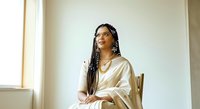 Young woman with long braided hair and glasses, wearing a cream-colored satin shirt, seated on a wooden chair against a neutral beige wall. Suitable for lifestyle, professional, and portrait projects.