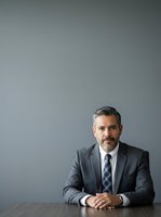 Middle-aged Caucasian male professional in formal business attire, seated at desk with hands clasped, wearing charcoal suit and plaid tie against neutral gray background for corporate portraits.