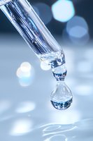 Crystal clear water drop forming from glass pipette against soft blue bokeh background, capturing moment of surface tension in laboratory setting.