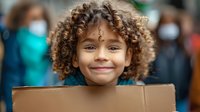 Young mixed-race girl with curly hair smiling while holding cardboard box during community volunteer event with masked people in background.