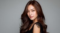 Young Asian woman with long wavy brown hair and natural makeup wearing black lace top poses confidently against gray background, professional beauty portrait.