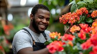 Happy African American male florist smiling while arranging colorful flowers in greenhouse nursery, professional gardener at work with blooming plants.
