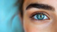 Close-up of striking blue eye with long eyelashes and defined eyebrow against turquoise background, showcasing natural beauty in macro photography.