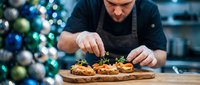 Professional chef garnishing gourmet appetizers on wooden board in festive kitchen with Christmas tree background for holiday catering and fine dining.