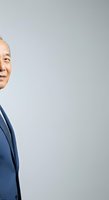 Middle-aged Asian male professional in navy business suit peering around corner against neutral background, suitable for corporate portraits and business communications.