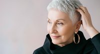 Senior woman with short white hair and gold hoop earrings wearing black turtleneck, touching her head with hand against neutral background. Mature female portrait for beauty, wellness, aging, and lifestyle content.