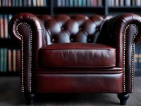 Luxurious burgundy leather armchair with rolled arms and silver nail head trim against blurred bookshelf background, showcasing classic Chesterfield design details.