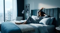 Man with brown hair and medium skin tone stretching in bed after waking, wearing casual clothing in modern bedroom with city skyline view, contemporary interior design.