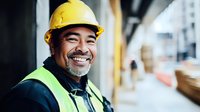 Cheerful Asian construction worker in yellow hard hat and safety vest smiling at construction site, showing positive workplace attitude and professionalism.