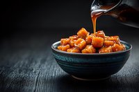 Caramel sauce being poured over golden caramel cubes in ceramic bowl on dark wooden surface for dessert preparation and confectionery cooking concepts.