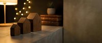 Three wooden miniature house figurines on a concrete surface with warm bokeh lights and books in a cozy dark interior setting.