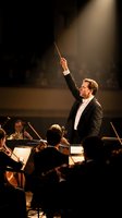 Professional orchestra conductor in formal tuxedo leading symphony performance with baton raised under dramatic stage lighting in concert hall.