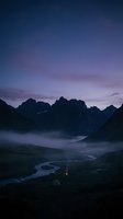 Dramatic mountain landscape at twilight with winding river through misty valley, purple and blue sky, jagged peaks silhouetted against dawn or dusk light. Perfect for travel and nature projects.