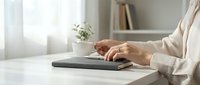 Woman hands typing on laptop computer at modern home office desk with plant and books in soft natural light for remote work and productivity.