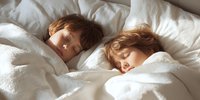 Two young children with auburn hair sleeping peacefully in white bedding, bathed in soft morning light.