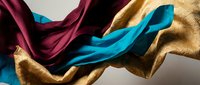 Luxurious silk fabric in vibrant burgundy, turquoise and gold colors flowing gracefully with elegant draping and smooth texture for fashion design projects.