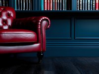 Luxurious red leather chesterfield armchair against dark blue wall with classic library bookshelf. Dramatic lighting highlights tufted upholstery and elegant curves.