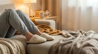 Cozy home relaxation scene with person in gray joggers and soft slippers resting on sofa with warm knit blankets and glowing lamp.