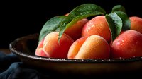 Ripe apricots with green leaves arranged in a dark ceramic bowl against black background, suitable for food photography, culinary content, and recipe illustrations.
