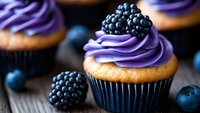 Fresh vanilla cupcake topped with vibrant purple buttercream frosting and garnished with ripe blackberries and blueberries on rustic wooden surface.