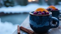 Fresh cranberries and orange slices in dark blue ceramic cup with cinnamon sticks on wooden surface against snowy winter background, cozy holiday mood.