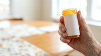Senior adult hand holding white prescription bottle with yellow cap in bright home interior setting for healthcare and medication concepts.