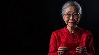 Senior Asian woman in elegant red lace dress holding traditional red envelope against dark background, celebrating lunar new year with warm smile and glasses.