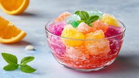 Colorful fruit jelly cubes in glass bowl with fresh mint leaves and orange slices on marble background for dessert recipes and food photography.