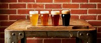 Four craft beer glasses with different colored ales and stouts displayed on rustic wooden barrel table against red brick wall background for brewery, pub, or beverage marketing.