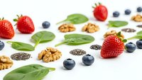 Fresh superfood ingredients scattered on white background with strawberries, blueberries, walnuts, chia seeds and spinach leaves for healthy eating.