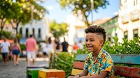 Young boy with dark skin tone and styled hair wearing a colorful patterned shirt, sitting on a bench in a vibrant town square surrounded by greenery, blurred pedestrians and colonial architecture in background.