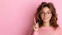 Young Caucasian woman with wavy brown hair and glasses pointing up with index finger, wearing pink sweater against pink background, showing excited expression.
