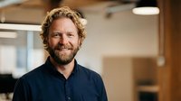 Smiling middle-aged Caucasian businessman with curly hair and beard in navy shirt standing in modern office workspace for corporate headshot photography.