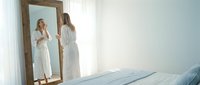 Woman in white dress looking at mirror reflection in bright bedroom with natural light from window.